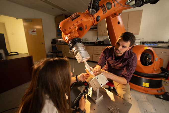 Two people sit to either side of a model of the human knee that is being grasped by a large orange robotic arm